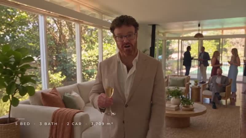 The host raises a glass in the sunroom while guests talk by the windows.
