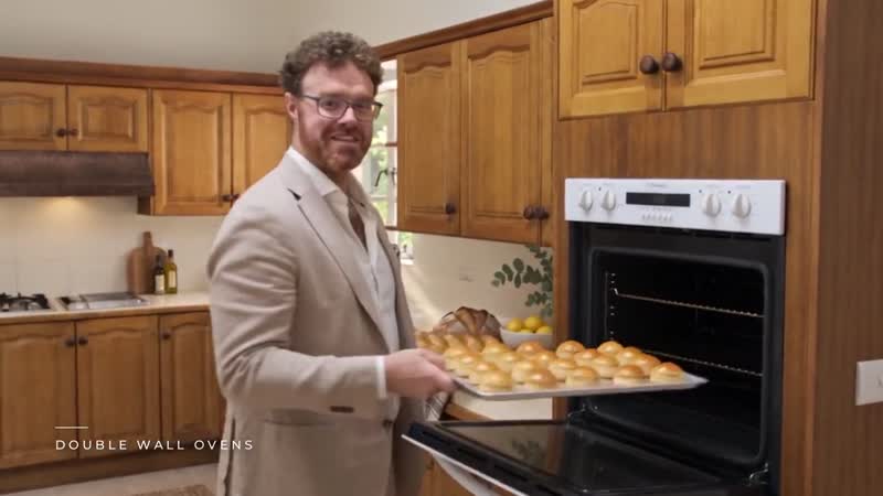 The host pulls a tray of bread rolls from the top wall oven in a timber kitchen.